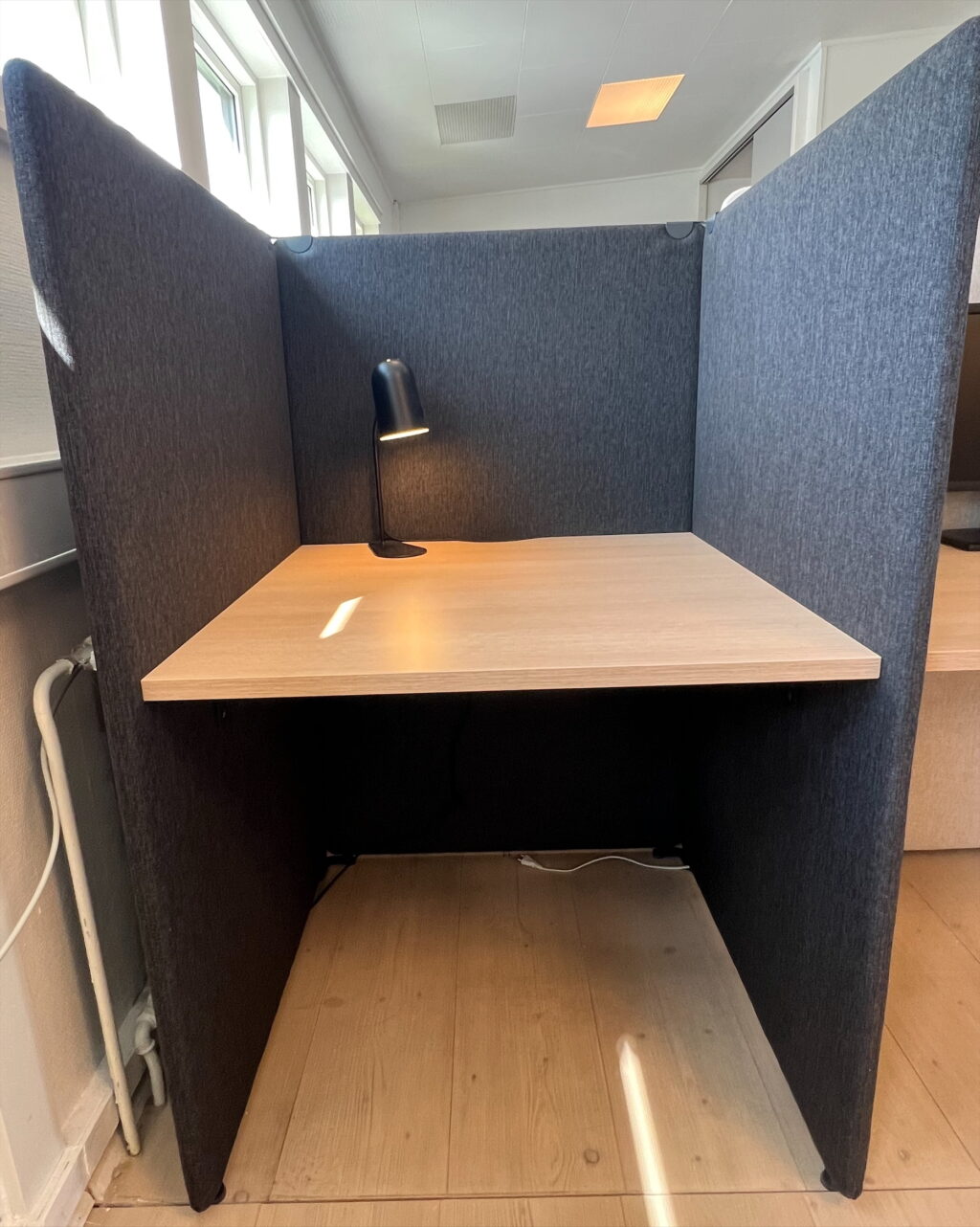 En enkel, afskærmet arbejdsniche med mørkegrå stofpaneler omkring et lyst træbord. En sort skrivebordslampe kaster varmt lys på bordfladen. Naturligt dagslys kommer fra vinduer til venstre, og lyse trægulvbrædder ses under. Loft med indbyggede lys og en afskærmet, rolig atmosfære uden personlige genstande.